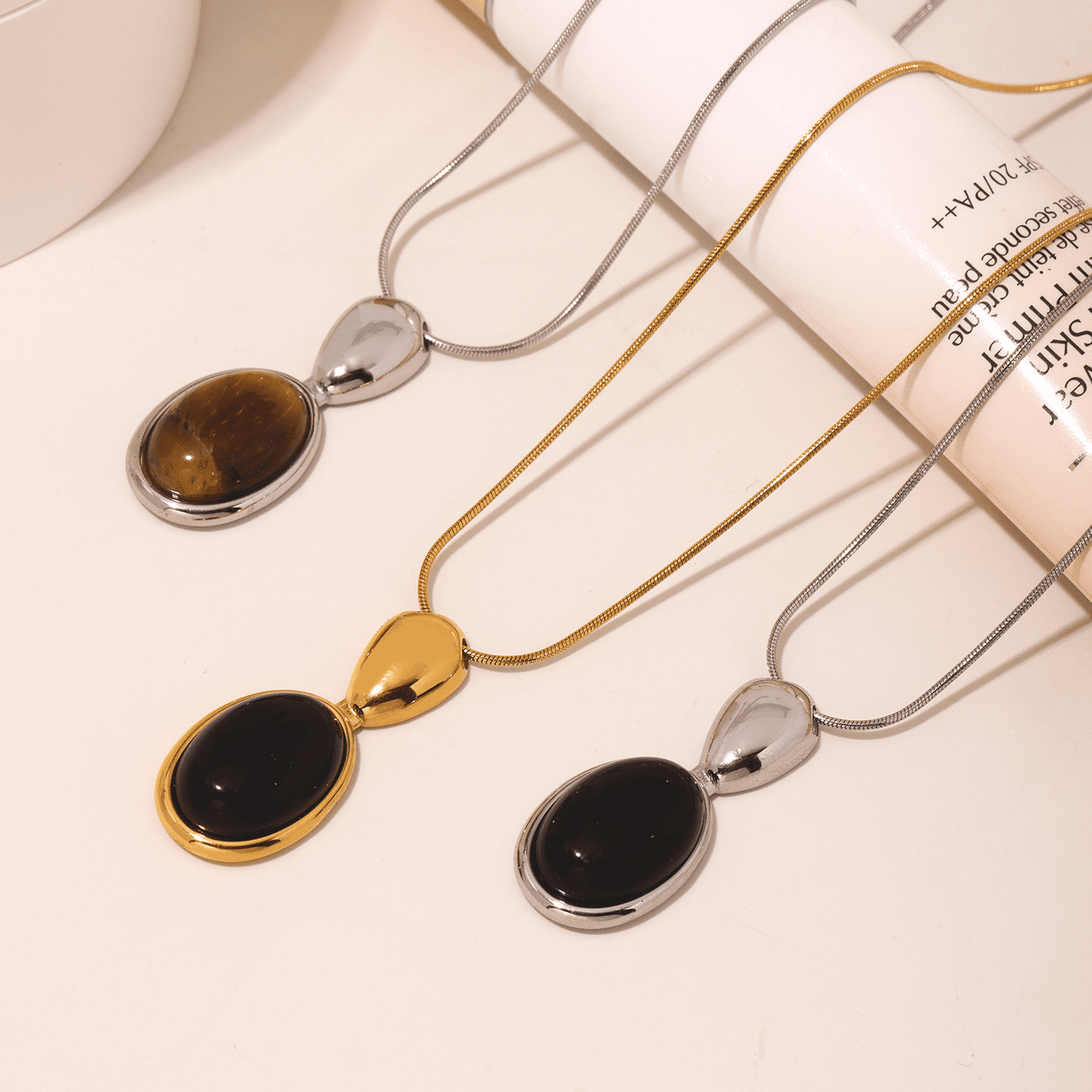 Two lariat necklaces with gold and silver color accents, featuring black oval pendants, displayed against a white background with textural elements.